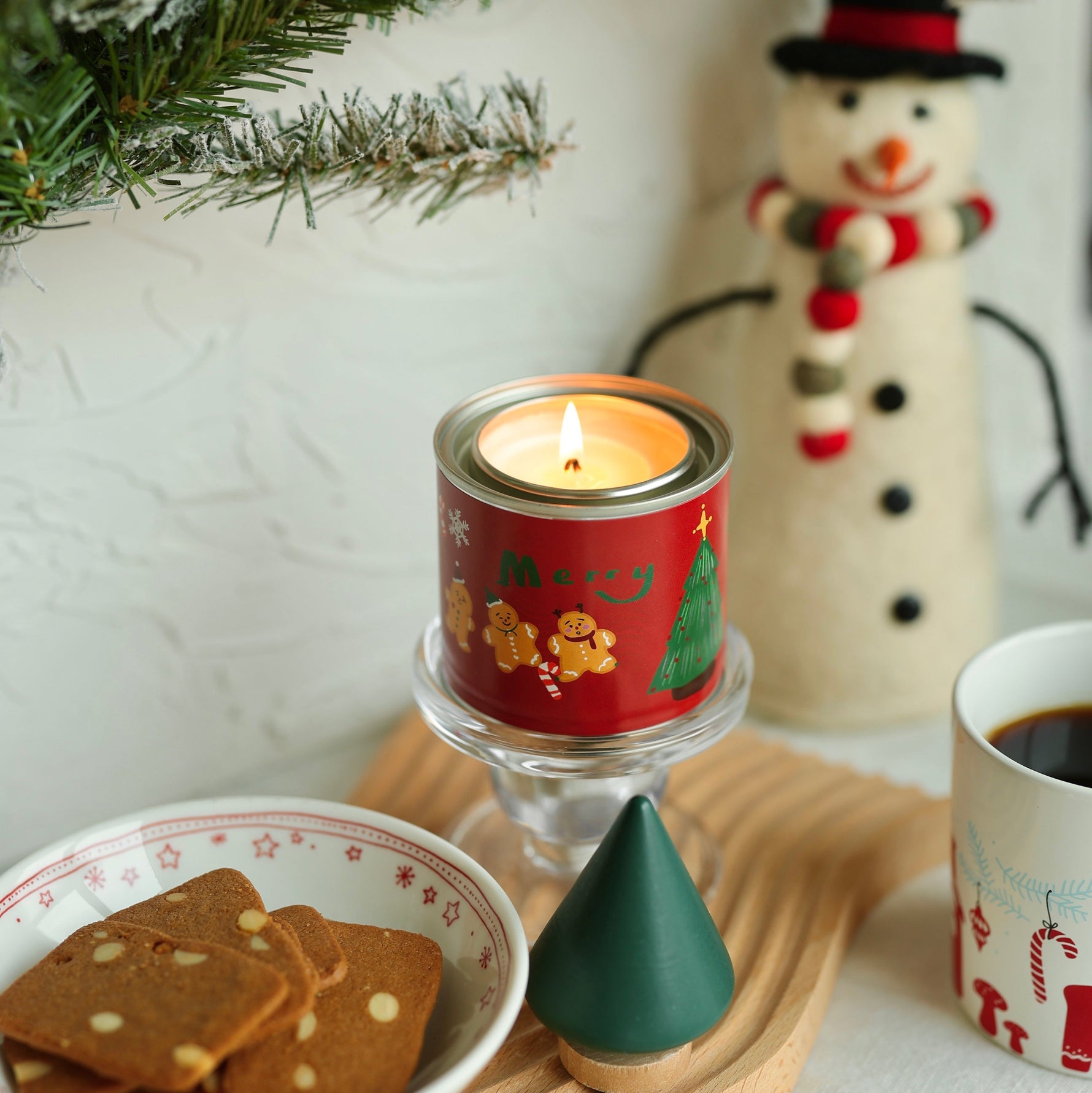 Candle in a red holder with cookies and a mug on a table, snowman decoration in the background.
