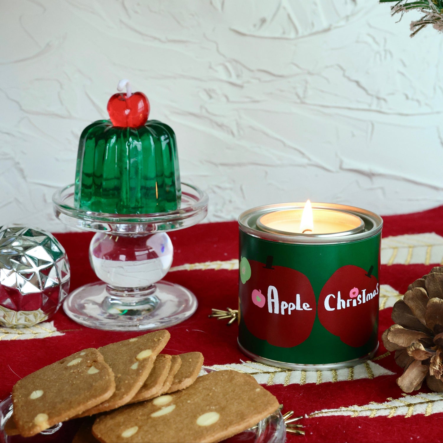 Christmas-themed candle with apple design, cookies, and green jelly dessert on a red tablecloth.